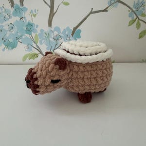 Puede incluir: Una figura de capibara tejida a ganchillo con hilo marrón claro y detalles en marrón oscuro en la nariz y las patas. El capibara tiene una pieza blanca, circular y enrollada sobre su espalda, que parece un cojín pequeño o una toalla.