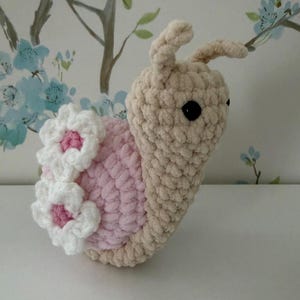 Puede incluir: Un juguete de caracol tejido a ganchillo. El cuerpo es beige, la concha rosa y adornada con flores blancas con centros rosas. Tiene ojos negros y antenas. El juguete está hecho de hilo suave y texturizado.