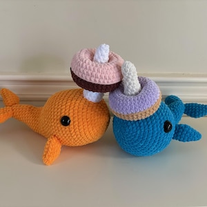 May include: Two crocheted stuffed narwhals, one orange and one blue, with a pink and brown crocheted donut on top of the orange narwhal.