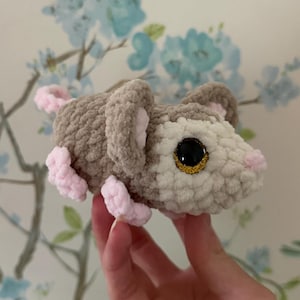 May include: A small, handmade crochet plush toy shaped like a mouse. The body is crafted from soft, textured yarn in shades of light brown and cream. It features large, round, dark brown plastic eyes with gold flecks, and small, light pink crocheted paws and a tail.