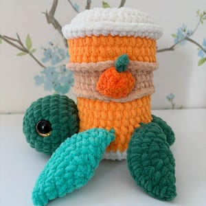 May include: A handmade crochet coffee cup cozy in orange, white, and tan, with a small orange pumpkin applique. Includes a green crochet ball with a black eye and green and teal crochet leaves. The cozy is designed to resemble a pumpkin spice latte.