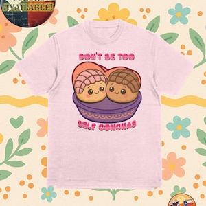May include: Pink t-shirt with a cartoon image of two concha pastries in a bowl. The text "Don't be too self conchas" is printed on the shirt.
