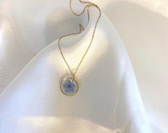 Puede incluir: Un collar de cadena dorada con un colgante redondo con una flor azul prensada.