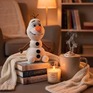 Peut inclure: Une figurine de bonhomme de neige crochetée en blanc avec un nez en forme de carotte orange vif, trois boutons noirs et des bras en forme de brindilles marron. Elle est posée sur trois livres intitulés Winter Wonders, Winter Cabin et Cozy Cabin Tales.