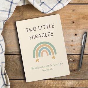 May include: A cream-colored journal with the text "TWO LITTLE MIRACLES" and a rainbow graphic. Below the rainbow is the text "MILESTONE AND PREGNANCY JOURNAL". A black pen rests on a wooden surface next to the journal.