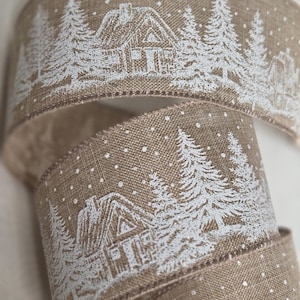 May include: A tan burlap ribbon featuring a white printed pattern of snow-covered houses and evergreen trees. The design includes small white dots representing falling snow across the textured fabric surface of the wide decorative ribbon.