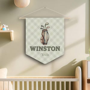 May include: A personalized wall banner featuring a golf bag with clubs on a light green checkered background. The banner displays the name "WINSTON" in gray letters, with "Keith" below it. The banner is suspended from a wooden dowel.