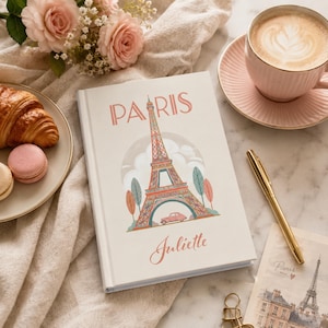 May include: Cream-colored journal with a Parisian theme. The cover features the Eiffel Tower, a pink car, and the name "Paris" in coral lettering. The name "Juliette" is written in script below the illustration. A croissant, macarons, and a cup of coffee are also visible.