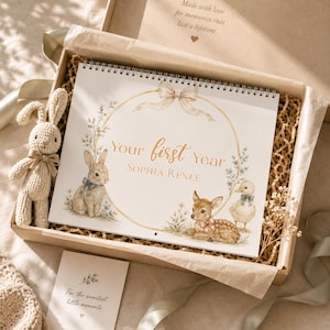 May include: A spiral-bound baby memory book titled Your First Year Sophia Renee, featuring illustrations of a rabbit, a fawn, and a duckling inside a gold circular frame with a bow. A small crocheted rabbit toy sits beside the book in a gift box.