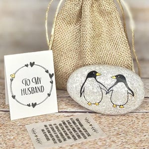 May include: A painted stone with two penguins holding hands, a small card that says "TO MY HUSBAND", and a burlap gift bag. The penguins are black and white with yellow feet and beaks. The card has a heart border.