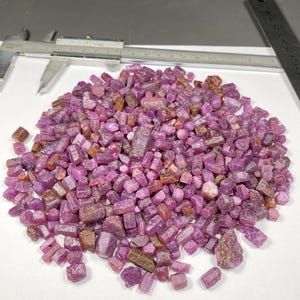 Könnte beinhalten: Ein Haufen roher, ungeschliffener Rubin-Edelsteine in verschiedenen Rosa- und Lilatönen. Die rohen Steine sind auf einer weißen Oberfläche verstreut, mit einem Metallmessschieber und Lineal im Hintergrund. Die Edelsteine haben ein natürliches, unpoliertes Aussehen.