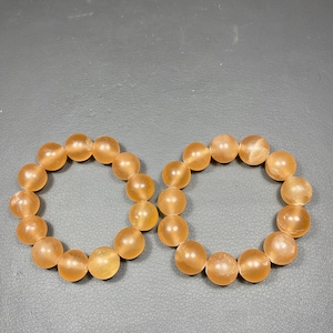 May include: Two circular bracelets composed of smooth, round, translucent orange beads. Each bracelet features a series of uniform, polished spherical stones strung together in a continuous loop, displaying a warm, soft, and glowing amber hue.
