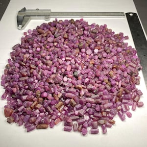 Peut inclure: Un tas de petits rubis bruts de différentes nuances de rose et de rouge. Un pied à coulisse en métal et une règle sont placés sur une surface blanche près des pierres précieuses. Les pierres sont éparpillées.