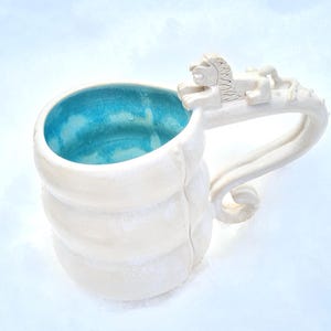 May include: White ceramic mug with a light blue interior. The mug has a sculpted handle with a lion figure. The body of the mug has a ribbed design. A unique and artistic piece for enjoying beverages.