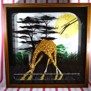 May include: A framed wall clock featuring a giraffe drinking water, with a yellow sun-shaped clock face. The clock has a brown wooden frame and a black background with silhouettes of trees and grass. The giraffe has a brown and tan pattern.