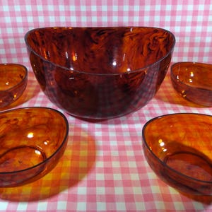 May include: A set of five amber-colored glass bowls. The set includes a large serving bowl and four smaller individual bowls. The bowls have a marbled appearance and are arranged on a pink and white checkered surface.