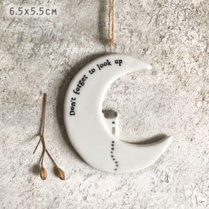 May include: White ceramic crescent moon ornament with the text "Don't forget to look up." The ornament is 6.5 cm x 5.5 cm and has a small house and dotted line design. A natural twine loop is attached for hanging.