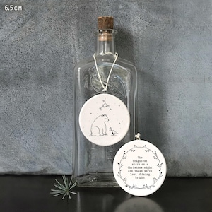 May include: Two white ceramic Christmas ornaments, each 6.5 cm in diameter, hang in front of a clear glass bottle. One features a bear and bird illustration, the other has text within a wreath: "The brightest stars on a Christmas night are those we've lost shining bright."
