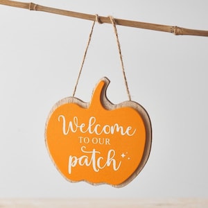May include: A pumpkin-shaped wooden sign with an orange centre and a light brown border. The sign reads "Welcome to our patch" in white script. It is suspended by a twine string from a bamboo rod.