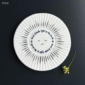 May include: A white ceramic plate measuring 10 cm in diameter with a sunburst pattern of radiating lines. The centre features a simple smiling face and the text "Good friends light up the world" written in a circular pattern around the face.