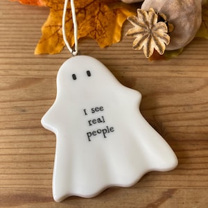 May include: A white ghost-shaped ornament with the text "I see real people" in black. The ghost has two black dot eyes and a white string for hanging. The background includes autumn leaves and seed pods.