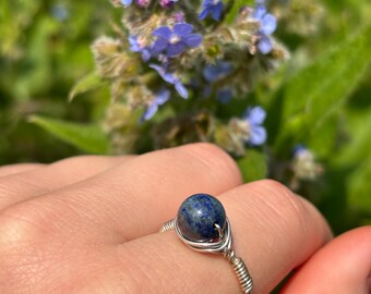 May include: A silver wire-wrapped ring featuring a round, deep blue gemstone. The ring is displayed on a hand, with a blurred background of green foliage and small blue flowers.