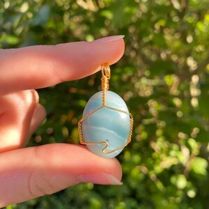 May include: A light blue oval gemstone pendant wrapped in gold-colored wire. The gemstone has a marbled appearance and is suspended from a gold wire loop. The background is a blurred green.
