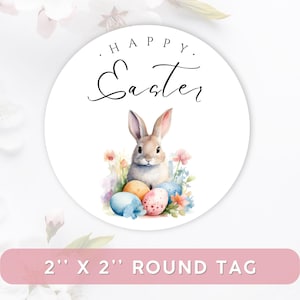 Op de afbeelding: Een ronde witte tag met de tekst "HAPPY Easter" in zwart script. Een aquarel illustratie van een konijn, kleurrijke eieren en bloemen staat in het midden. De tag is 5,08 cm x 5,08 cm groot.