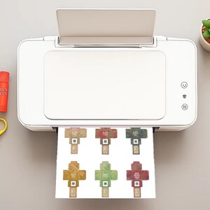 Pode incluir: Uma impressora branca imprime designs de embalagens de produtos em forma de cruz em v&aacute;rias cores. Ao lado da impressora est&atilde;o tesouras amarelas, cola e uma pequena planta suculenta em vaso. Os desenhos s&atilde;o impressos em papel branco.