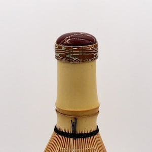 Op de afbeelding: Een bamboe matcha klopper met een donkerrode en goudkleurige dop. De klopper heeft een natuurlijke bamboe handgreep en een zwarte draadbinding. De dop heeft een gemarmerd ontwerp. De klopper wordt gebruikt voor het bereiden van matcha thee.
