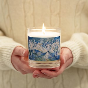 May include: A white candle with a glass jar and a label featuring a painting of white swans on a blue lake. The candle is lit and the flame is visible.