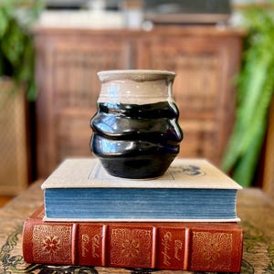 Könnte beinhalten: Eine handgefertigte Keramiktasse mit einem strukturierten, wellenförmigen Design. Die Tasse hat ein zweifarbiges Farbschema mit einem beigen Rand und einem schwarzen Körper. Sie steht auf zwei antiken Büchern mit blauen und roten Einbänden.