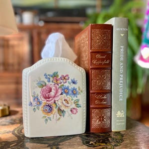 Puede incluir: Una funda de cerámica color crema para caja de pañuelos con un ramo de flores rosas, amarillas y azules. Junto a ella hay dos libros de tapa dura: un ejemplar de David Copperfield de Charles Dickens en piel marrón y Pride and Prejudice de Austen.