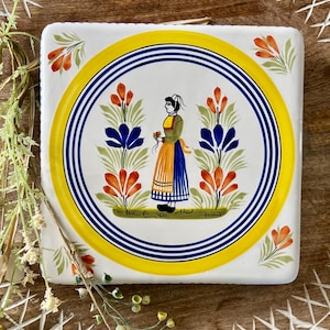 May include: Square ceramic tile with a hand-painted design. The tile features a central circular image of a woman in traditional attire, surrounded by orange and blue floral motifs. The border has yellow and blue concentric circles.