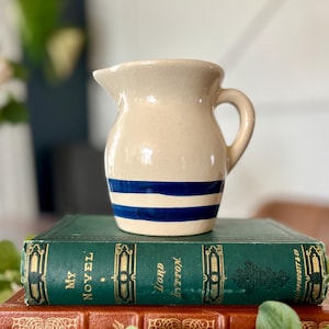 May include: A cream-coloured ceramic jug with a handle and spout, decorated with two blue horizontal stripes. It rests on a stack of antique books with green and brown covers and gold lettering.