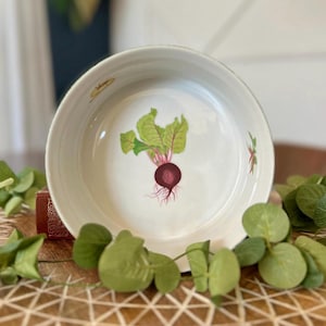 Może przedstawiać: Biała ceramiczna miska z namalowaną ilustracją buraka. Burak ma ciemnoczerwony korzeń, zielone liście i różowe łodygi. Misa otoczona jest zielonymi liśćmi.