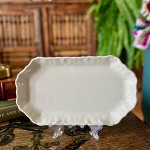 Puede incluir: Una bandeja de servir rectangular de cerámica en color blanco roto con un borde festoneado y ondulado. La bandeja presenta un acabado liso y brillante y se muestra sobre un soporte de plástico transparente, destacando su forma alargada.