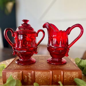 Può includere: Una zuccheriera in vetro rosso con coperchio e una lattiera abbinata. Entrambi i pezzi hanno manici ornati e poggiano su un libro marrone con scritte dorate. Il set è esposto su una superficie di legno.