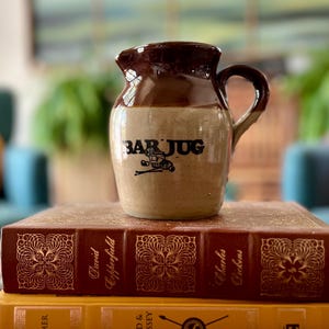May include: A ceramic jug with a two-tone brown and tan glaze, featuring a handle and a spout. The text "BAR JUG" is printed in black on the front, accompanied by a small graphic of a cocktail glass and stirrers. The jug rests on two books.
