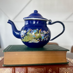 May include: A cobalt blue enamel teapot with a peacock design. The teapot has a black handle and a lid. The peacock design features colorful flowers and birds. The teapot is sitting on top of a green book and a brown book.