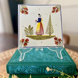 May include: Square ceramic tile with a hand-painted design of a woman in traditional clothing holding flowers. The tile has a white background with red, green, and blue accents. The tile is displayed on a clear acrylic stand, resting on a stack of green books.