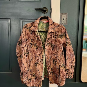 Könnte beinhalten: Eine langärmelige Knopfjacke aus strukturiertem Stoff im Wandteppich-Stil mit einem dichten Blumen- und Pflanzenmuster in Altrosa, Braun und gedecktem Grün. Das Innenfutter zeigt ein kontrastierendes Muster in Grün und Gold.