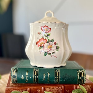 Op de afbeelding: Een crèmekleurige keramische pot met een deksel, versierd met een bloemenontwerp met rode en witte bloemen en groene bladeren. De pot staat op een stapel vintage boeken met groene en bruine covers.
