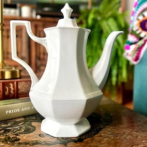 May include: A white porcelain teapot featuring an octagonal body, a matching lid with a pointed finial, a curved handle, and a tapered spout. The base and the shoulder of the teapot are decorated with a subtle beaded trim detail.
