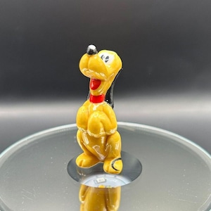 May include: A small ceramic figurine of Pluto, the Disney dog. Pluto is yellow with black ears and a red collar. He is standing on his hind legs with his front paws raised.