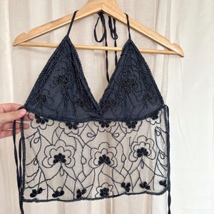 Peut inclure: Haut de bikini noir avec un motif floral. Le haut présente un corsage perlé et une section inférieure transparente brodée. Les bretelles sont fines et se nouent dans le cou. Le haut est suspendu à un cintre en bois.