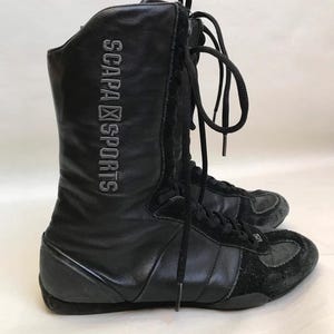 May include: Black leather and suede high-top boots with laces. The boots have the text "SCAPA SPORTS" printed on the side. The boots are designed for athletic activities.