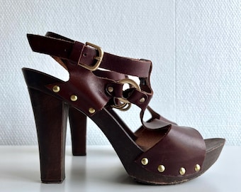 Könnte beinhalten: Braune Leder-Plateausandalen mit hohem Holzabsatz. Die Sandalen haben ein T-Riemen-Design mit goldfarbener Hardware und einem Schnallenverschluss. Die Plattform ist mit kleinen goldenen Nieten besetzt.