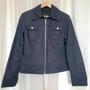 May include: A dark blue denim jacket with a silver zipper closure, two chest pockets with button closures, and a classic collar. The jacket is hanging on a wooden hanger.