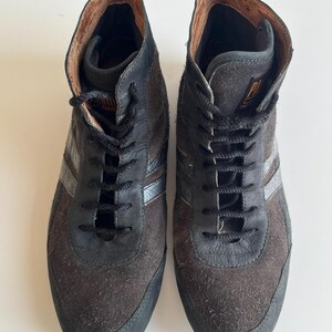 Pu&ograve; includere: Un paio di sneakers alte in pelle scamosciata marrone e pelle nera. Le sneakers hanno lacci neri, una striscia argentata e una suola nera. L'interno &egrave; di colore marrone chiaro.